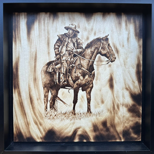 A framed wood burning artwork depicting a cowboy on horseback against a stylized, textured background.